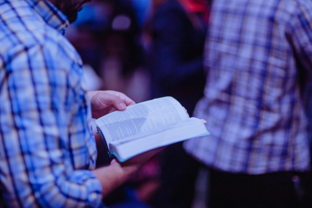 A man stands indoors reading a Bible, focusing amidst a crowd.