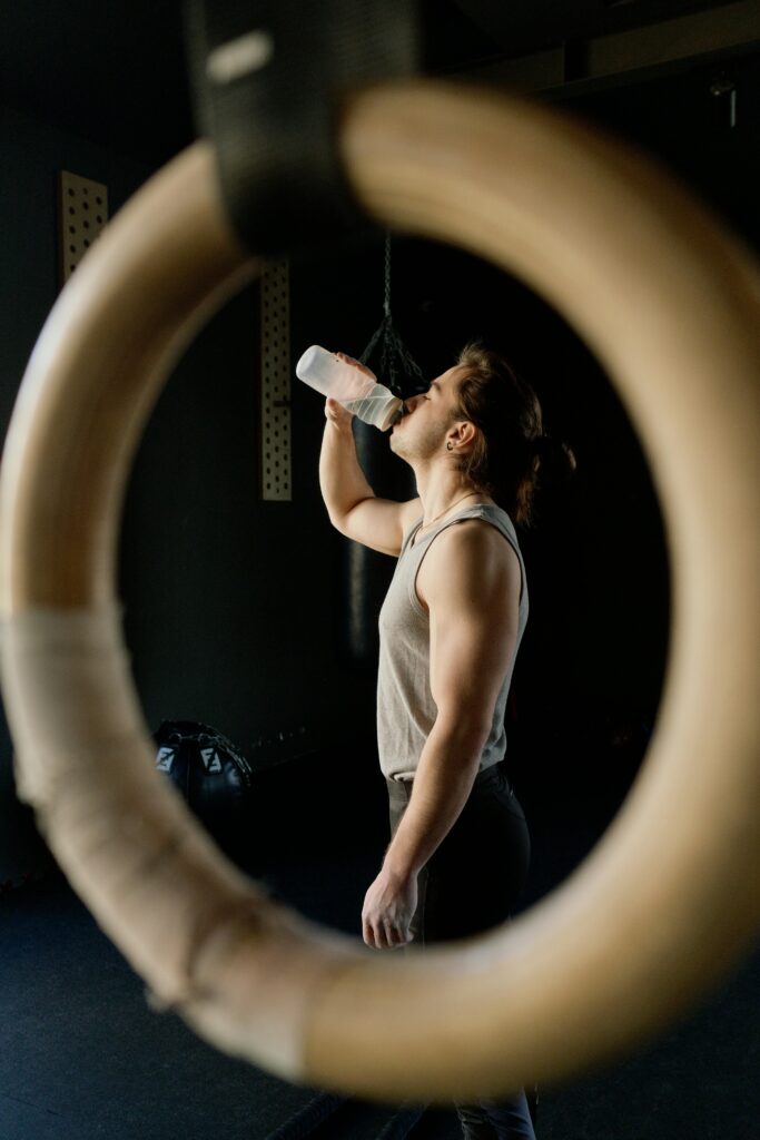 Caucasian man drinking water in a gym setting, framed by gymnastic rings.